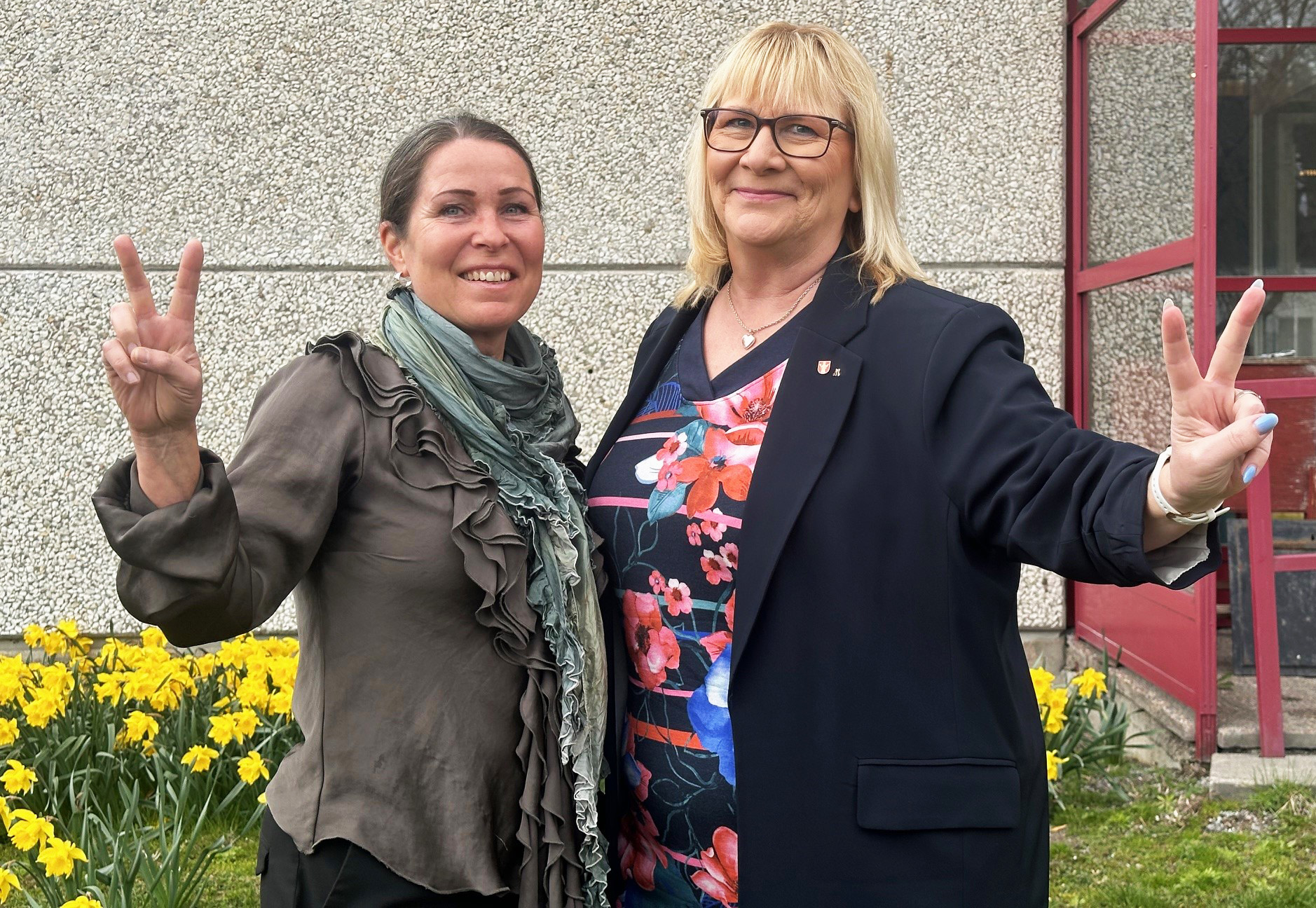 foto på samhällsbyggnadsdirektör Carina Molin och kommunstyreksens ordförande Stina Lundgren (M) som ser glada ut där de står framför kommunhuset. I rabbaten i bakgrunden syns påskliljor.