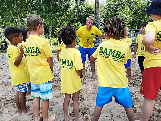 En grupp barn med gula fotbollkläder står i ring runt en vuxen tränare som ser ut att instruera barnen. 
