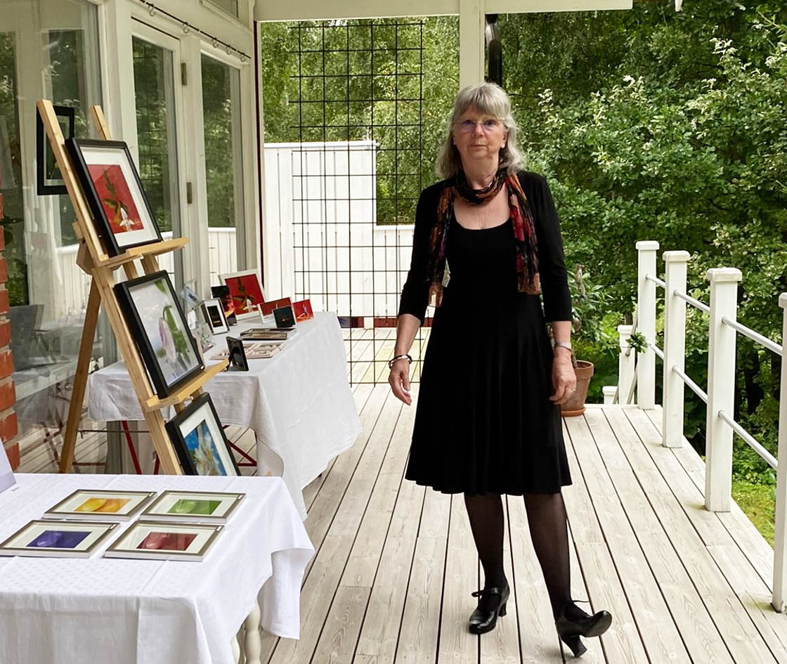 Berit Almér står på en veranda omgiven av konst. Bland annat visas en tavla på staffli och fler tavlor ligger på ett bord.