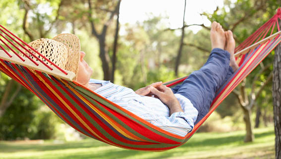 En man, med solhatt på huvudet, vilar i en randig hängmatta. Sommargrönska i bakgrunden.